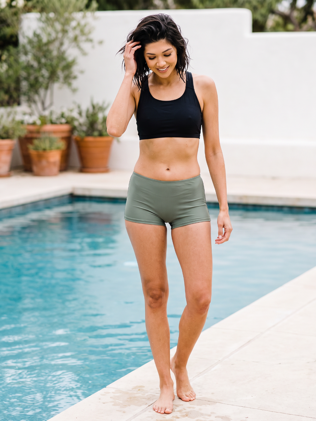 A woman wearing a black sports bra and olive green athletic shorts walks barefoot along the edge of a swimming pool.