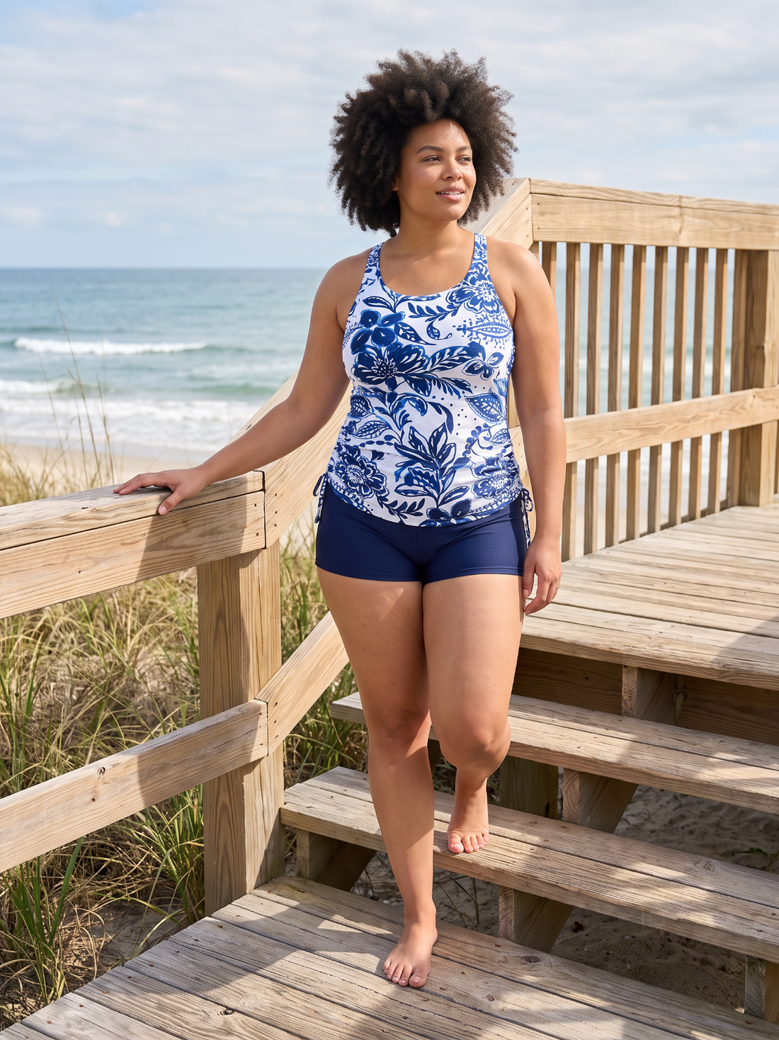 A woman with an afro hairstyle walks down wooden beach boardwalk stairs wearing a blue and white floral print tankini top and navy blue swim shorts, with the ocean and dunes in the background.
