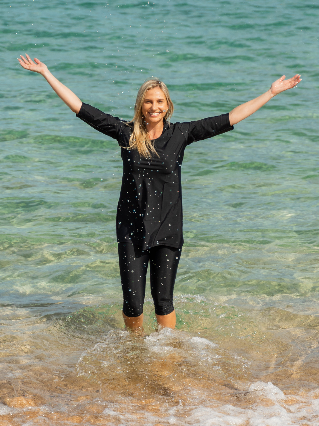 Woman wearing black Capri Swim Leggings and a matching swim top standing in shallow ocean water with arms raised