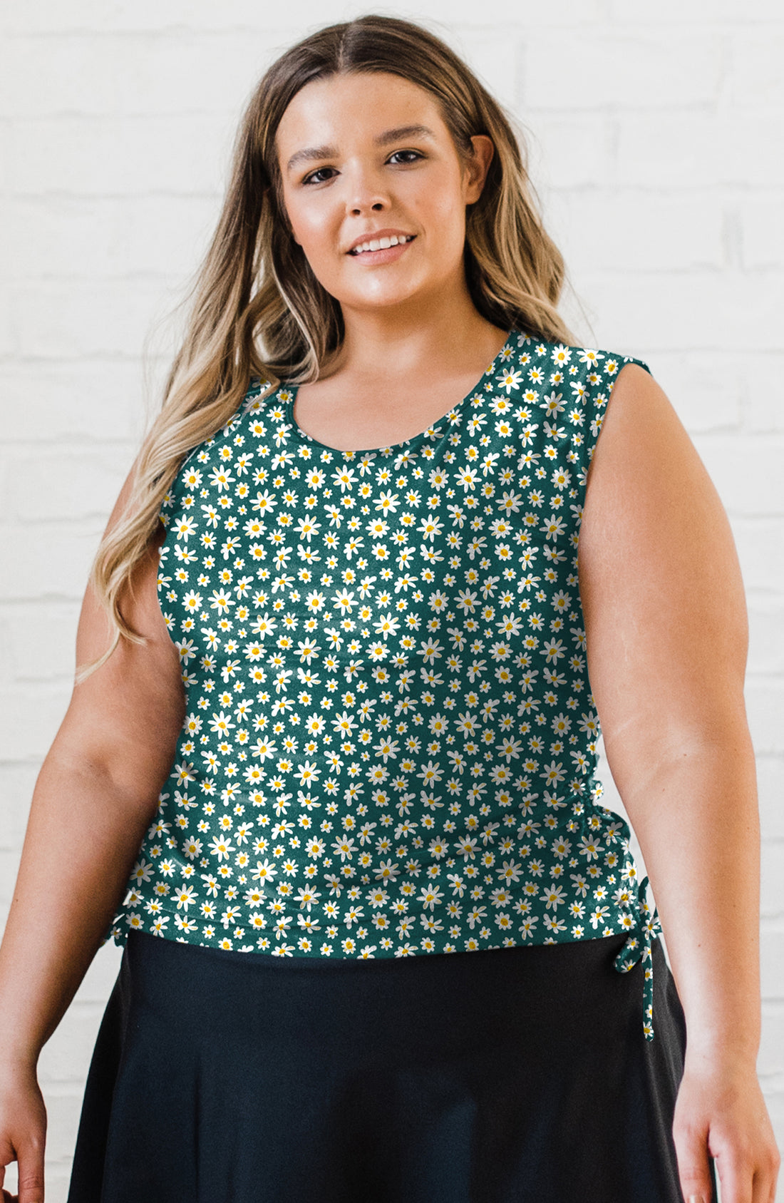 Woman wearing a green floral sleeveless top against a white brick wall.