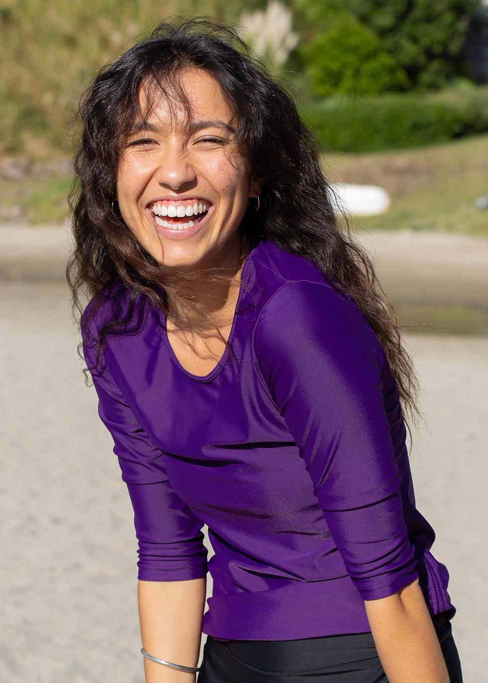 A person wearing a purple swim top with a round neckline and 3/4 sleeves, standing near a body of water.